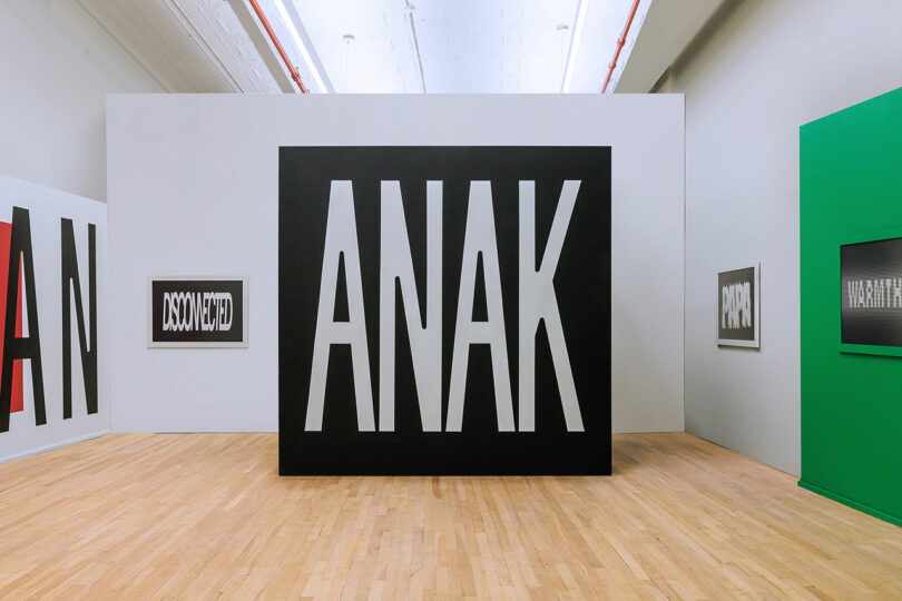 A gallery space with bold 'ANAK' typography on a central black box and framed text art on the walls.
