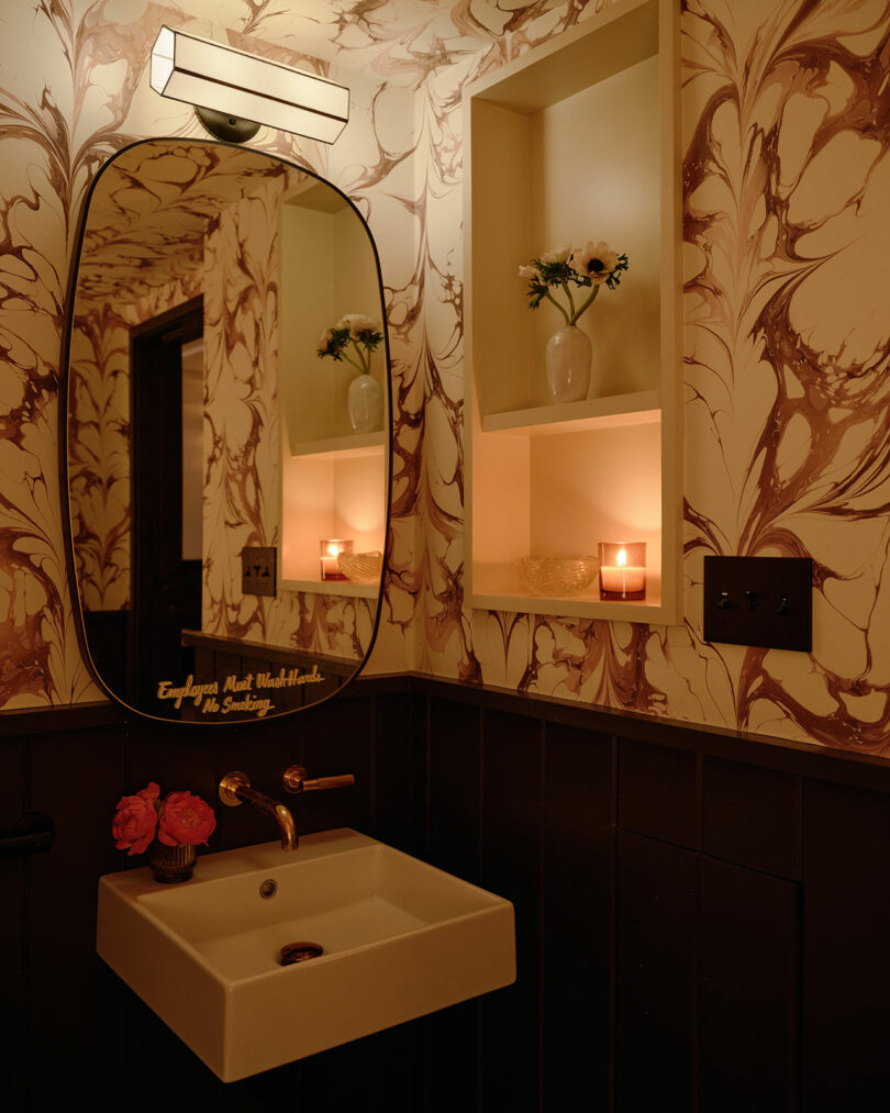A stylish bathroom with marbled wallpaper, an oval mirror, and candlelight.