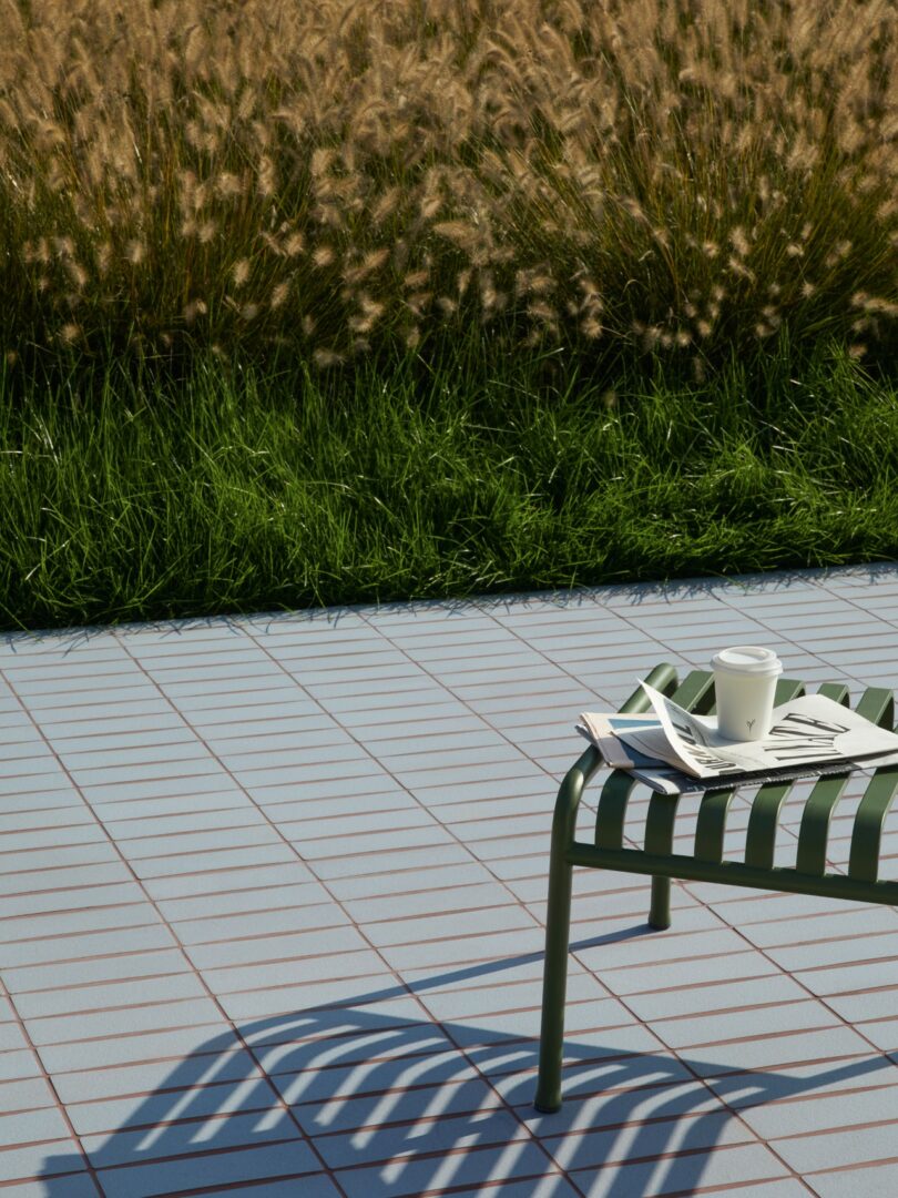 A green metal bench on a tiled outdoor patio with tall grass in the background.