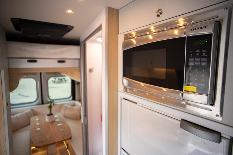 Close-up of a stainless steel microwave and refrigerator built into the van's cabinetry with a wooden table in the background.