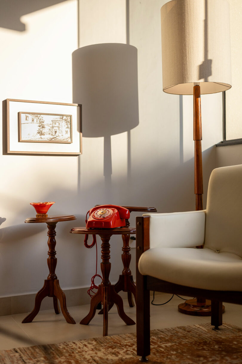 A vintage red rotary phone on a wooden side table next to a classic white armchair.