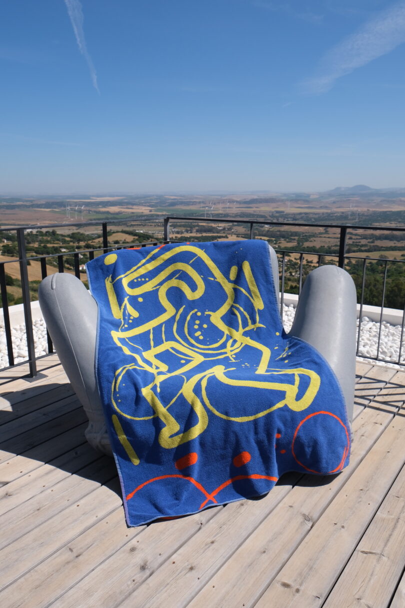 A modern lounge chair on a wooden deck featuring a blue and yellow abstract art towel overlooking a scenic landscape.