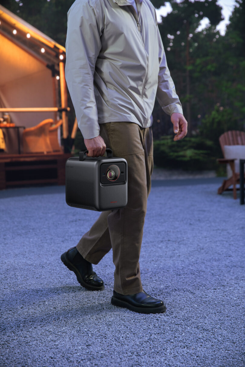 A person carrying the compact Anker Nebula projector by its handle outdoors on a gravel path.