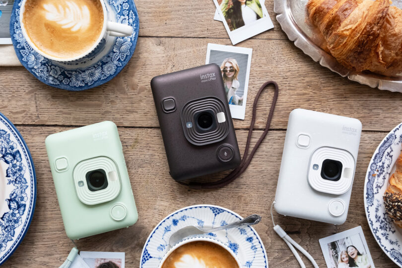 Three Instax Mini LiPlay cameras in green, bronze, and white on a wooden table with coffee and photos.