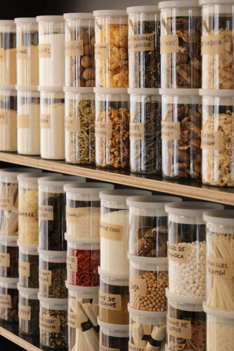 Labeled plastic containers filled with spices and grains on minimalist shelves.