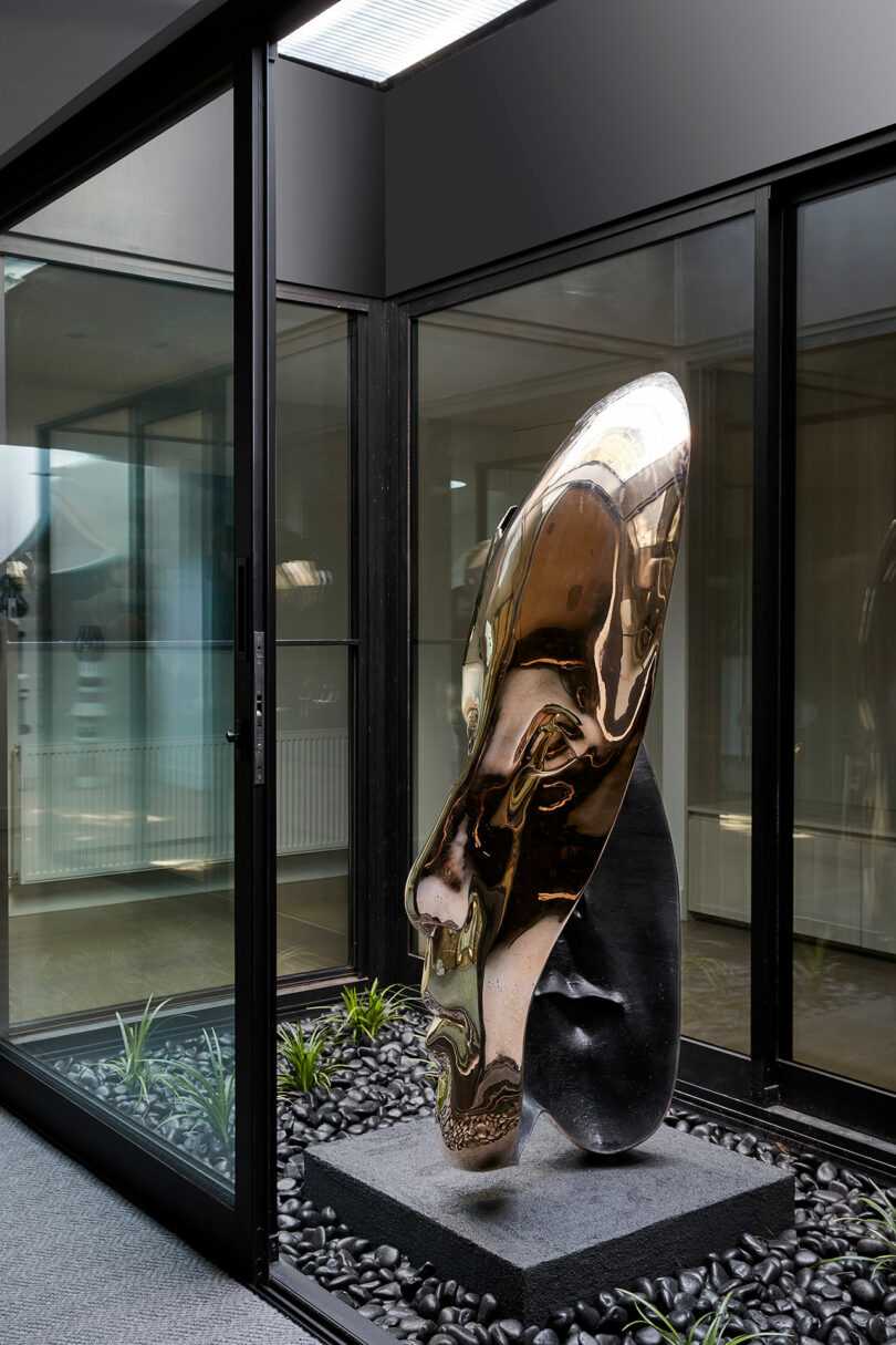 Elongated metallic sculpture on a stone base displayed in a glass-enclosed courtyard with black pebbles.
