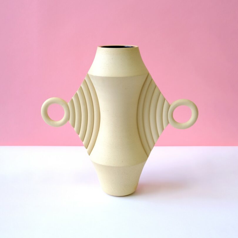 A beige geometric ceramic vase with circular handles against a soft pink background.