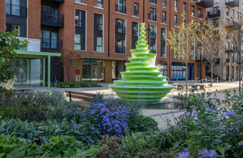 The lime green fountain situated in a modern urban courtyard with brick buildings and purple flower beds.