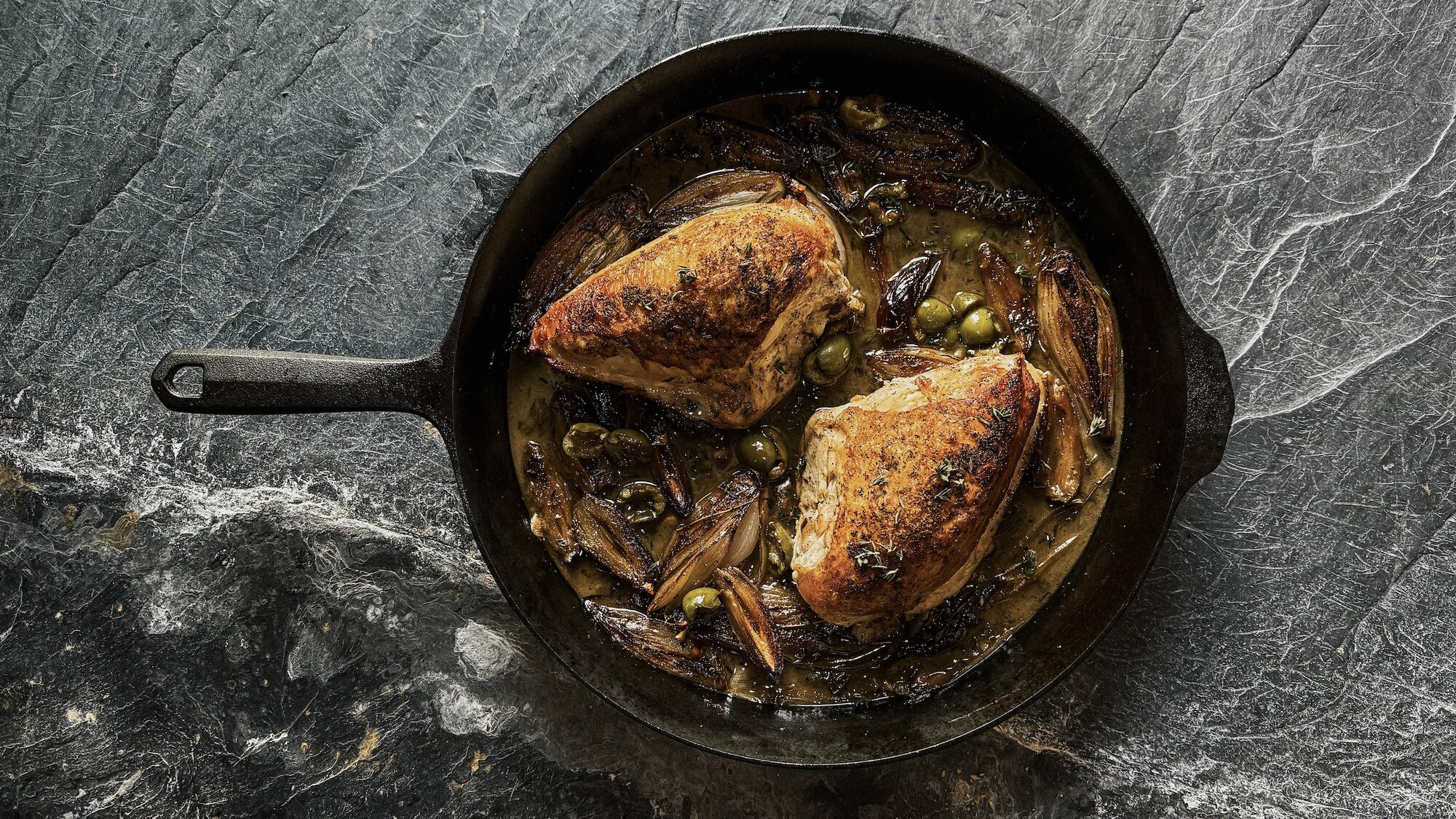 A close-up of golden brown pan-roasted chicken pieces garnished with green olives and dates.