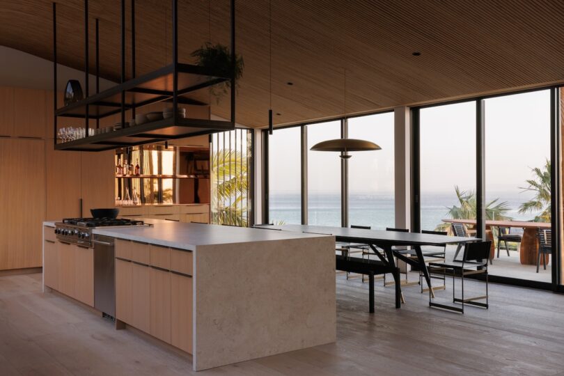 Modern kitchen with wood finishes, marble island, and large windows overlooking the ocean.