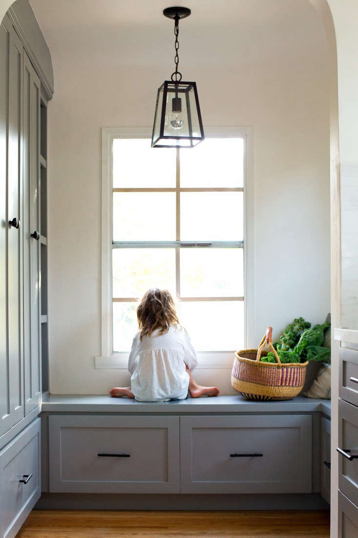 Child sitting on a gray Shaker-style window seat in a bright kitchen nook.