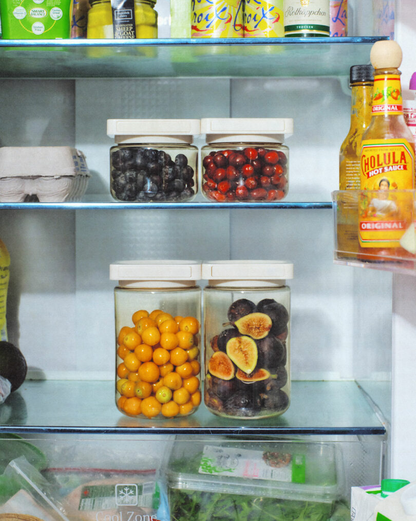 A modern kitchen refrigerator shelf displays organized glass jars with blueberries, cranberries, and dried figs.