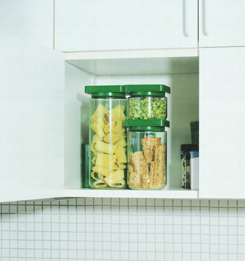 Modern kitchen cabinet with white cabinetry and tiled backsplash, featuring transparent storage jars with green lids holding pasta and snacks.