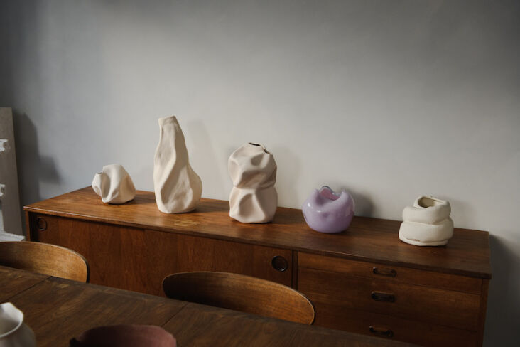 A wooden sideboard displaying abstract ceramic and glass vessels under soft light.