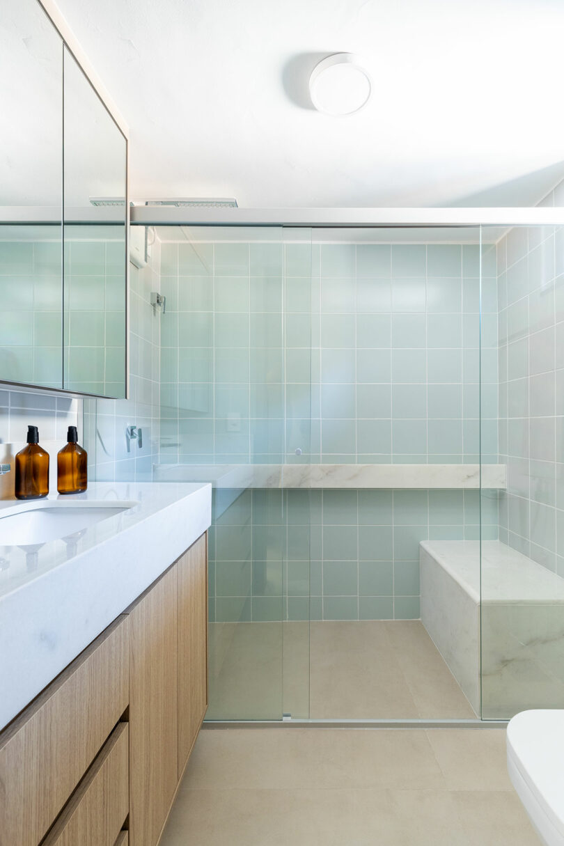 A modern bathroom with a glass-enclosed curbless shower, a built-in wooden bench, and amber soap dispensers.