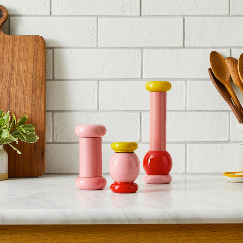Three modern salt and pepper grinders in vibrant pink and yellow finishes.