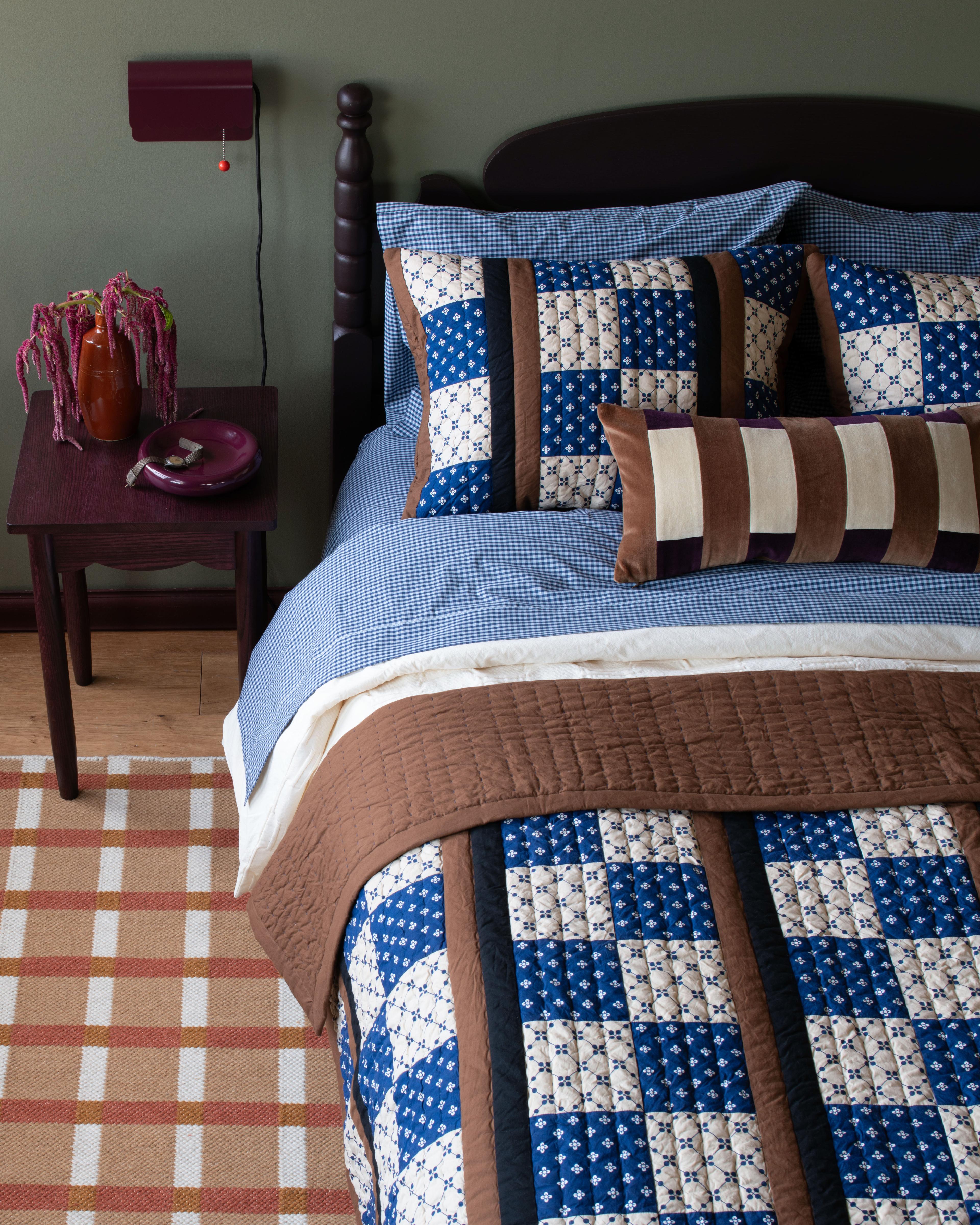 A bed layered with a thick comforter, a quilt, and a rug placed underneath for extra texture.