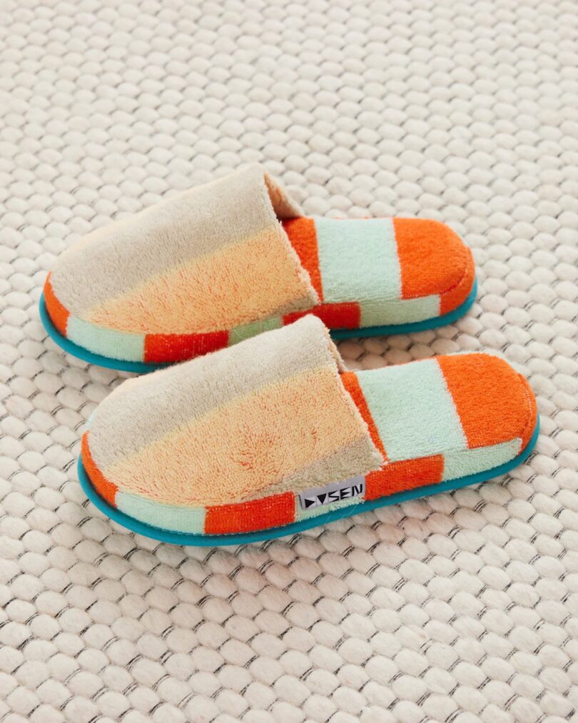 Orange and teal blue striped terry cloth house slippers on a light background.
