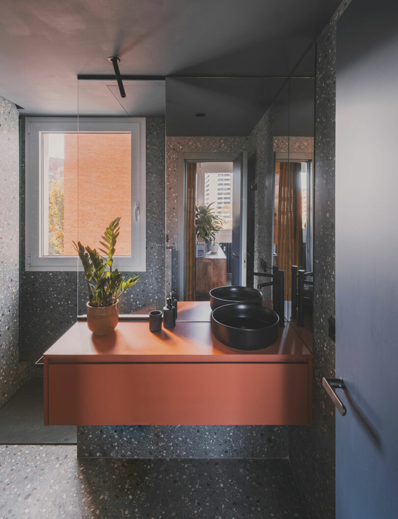 Modern bathroom with terrazzo walls, an orange vanity, and large mirrors that reflect light and space.