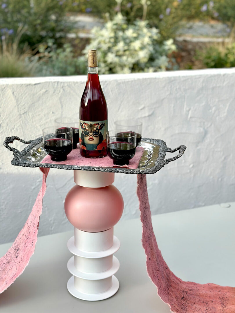 A wine display featuring a bottle and glasses on an ornate tray atop a sculptural pedestal with pink paper accents.