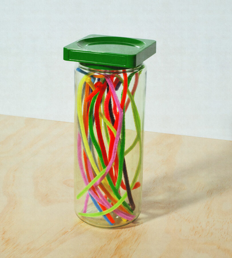 A clear glass jar with a green lid filled with colorful pipe cleaners sits on a light wood surface.