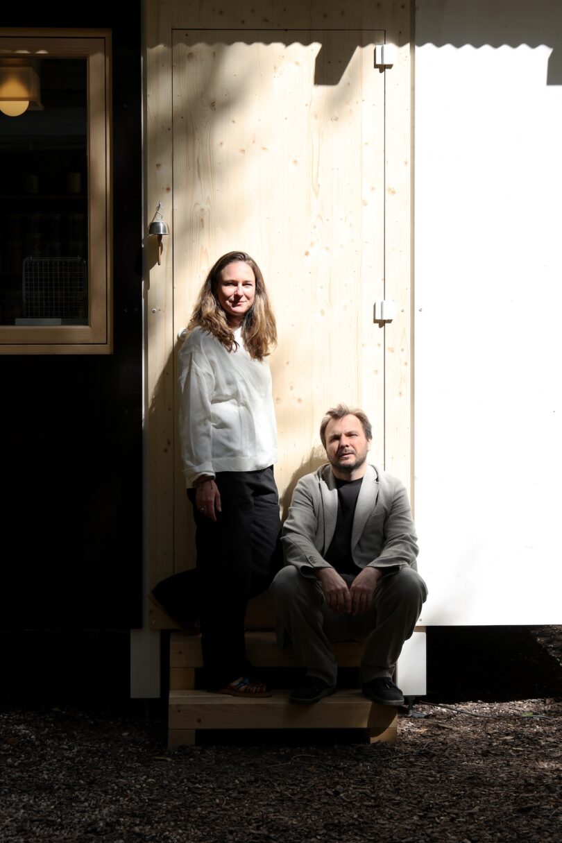 A man and woman sitting on the wooden steps of a modern micro-house in natural light.