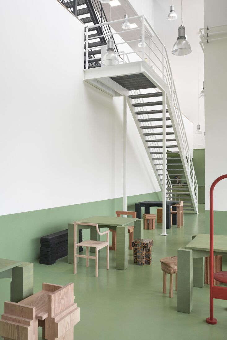 A wide industrial loft space with green floors and walls, featuring minimalist wooden furniture and a large metal staircase.