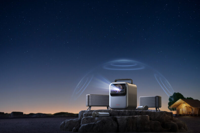 A portable projector setup outdoors at night projecting a bright image next to a tent and professional speakers.