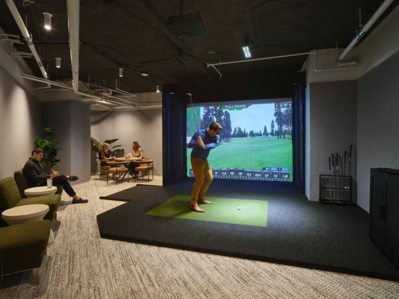 An industrial-style room featuring a large golf simulator screen and green turf.
