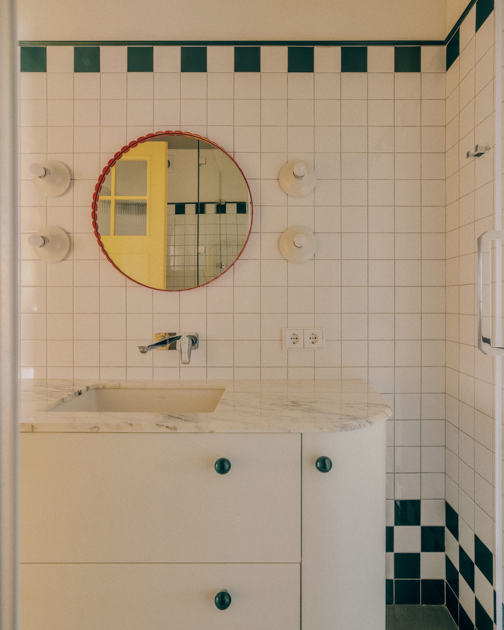 A modern bathroom featuring green checkerboard accents, a round red-framed mirror, and a marble countertop.