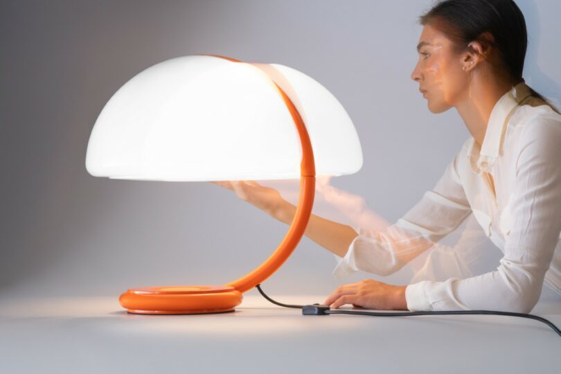 A woman adjusting the white dome shade of an orange Serpente table lamp to show its swivel movement.