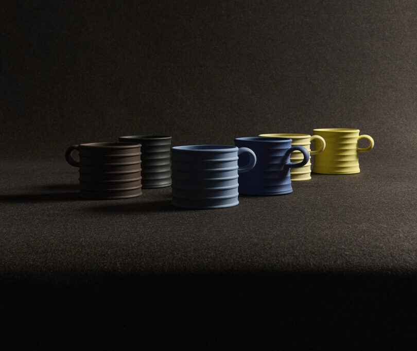 A lineup of five ribbed ceramic mugs in brown, black, blue, and yellow shades.
