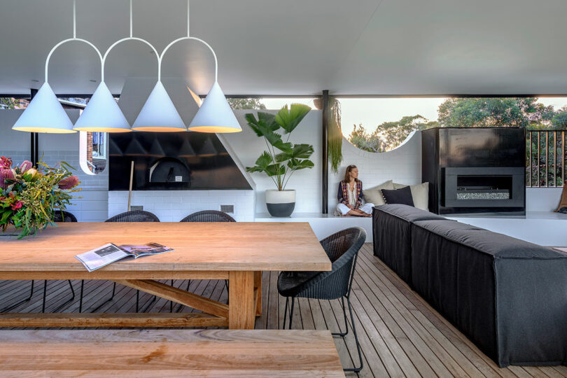 A woman relaxing on a black outdoor sofa next to a contemporary fireplace on a covered deck.