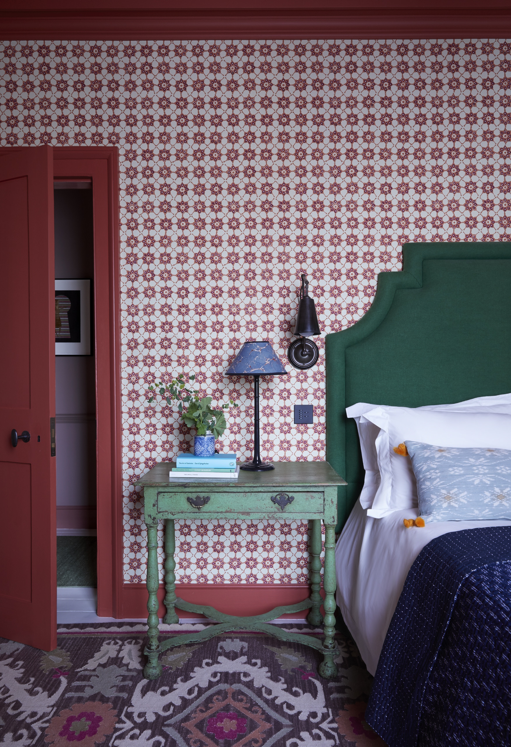 A small bedroom with red and pink patterned wallpaper and a deep juniper velvet upholstered bed.