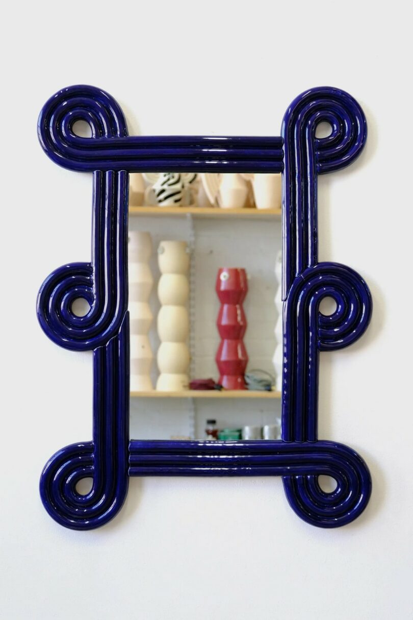 A rectangular mirror with a dark blue glossy sculptural coil frame on a white wall.
