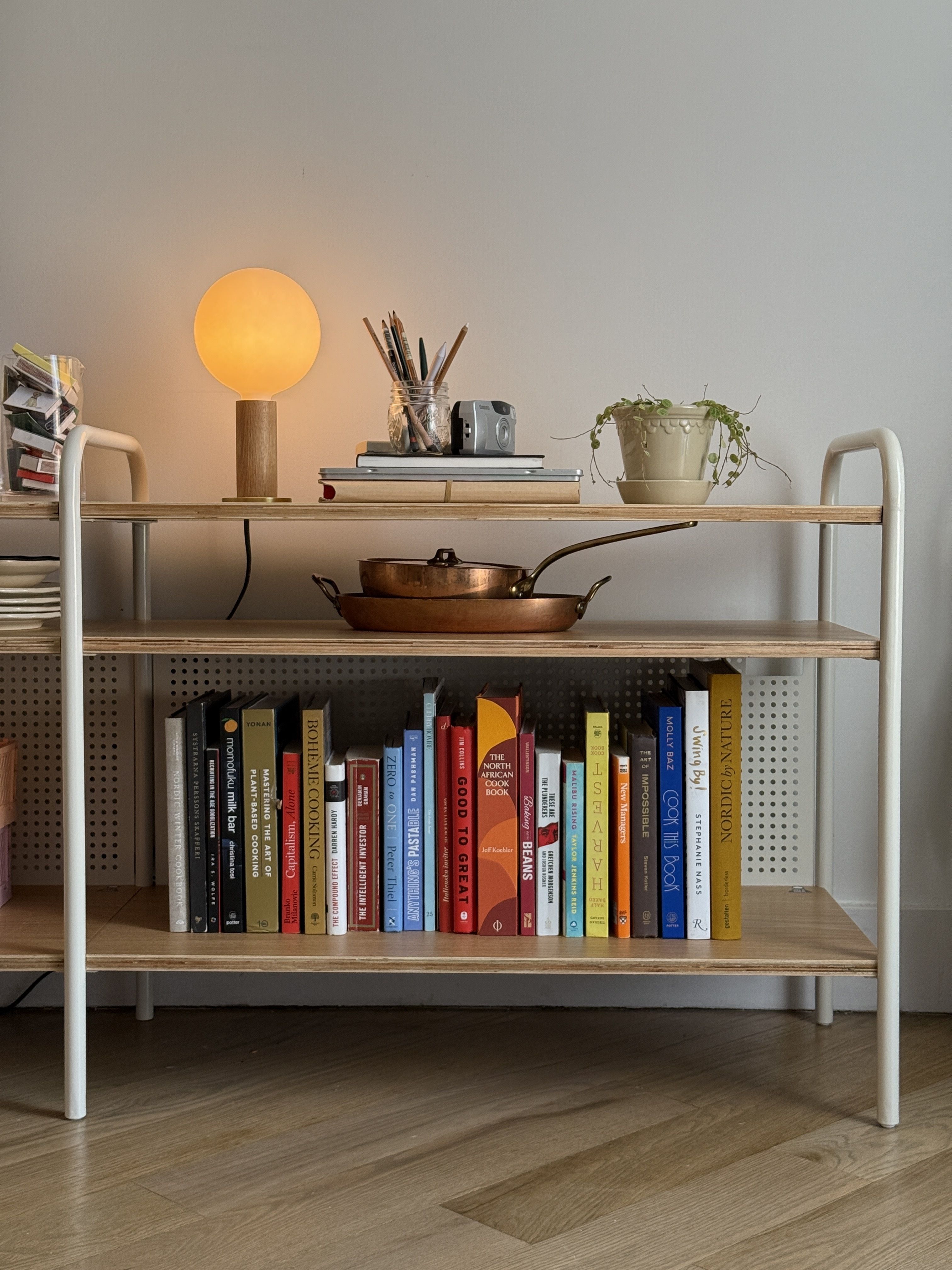 Close-up of a minimalist wooden shelf holding a variety of books, a warm lamp, and kitchenware against a neutral wall.