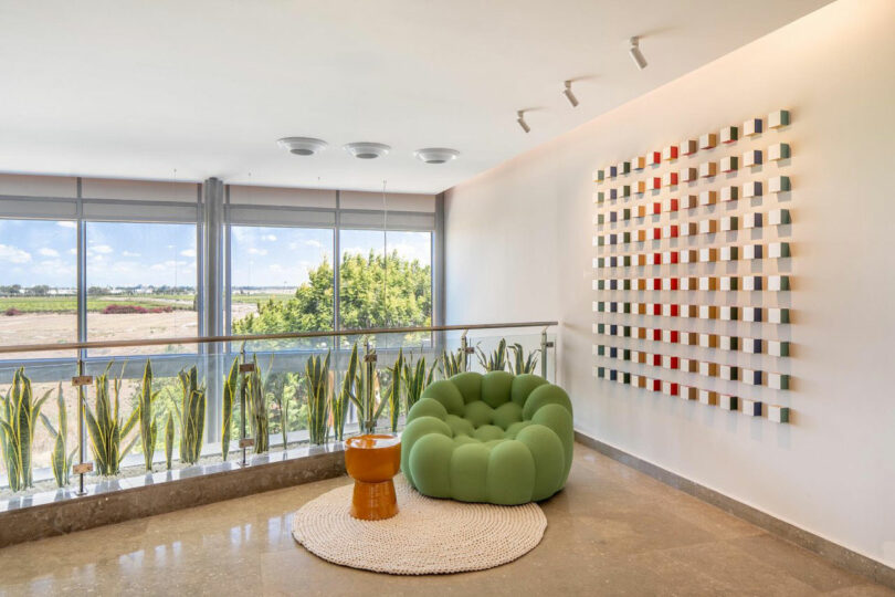 Lounge area with a green chair next to large windows overlooking an outdoor terrace with plants.