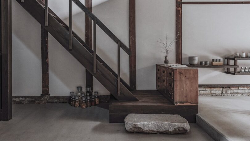 Minimalist interior with a rustic wooden staircase, a simple storage chest, and a single dried branch in a vase.