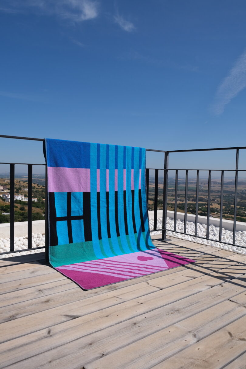 A geometric-patterned towel with bold blues and pinks hanging over a black metal railing with a scenic background.