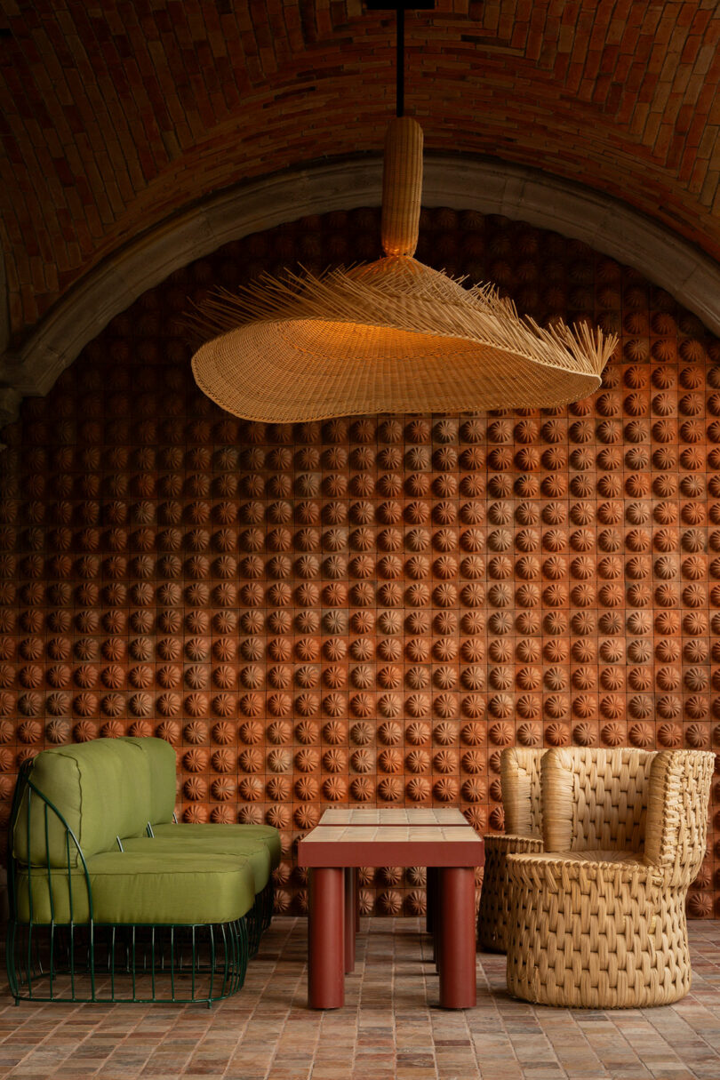 A large woven pendant lamp hanging over a green metal sofa and red table in a room with a brick arch.