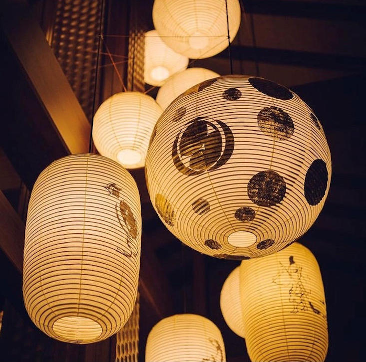A group of hanging Japanese lanterns with thick black circular outlines providing a warm orange glow against a dark ceiling.