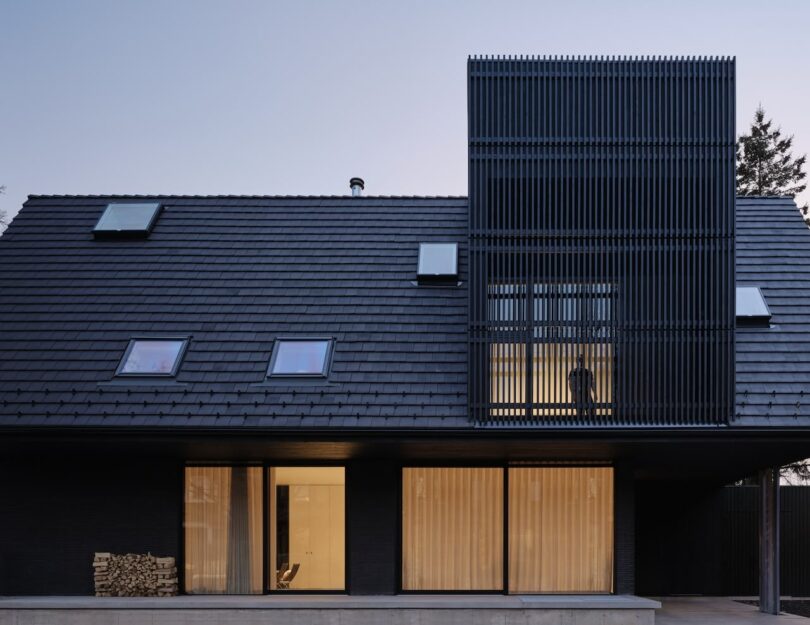 Modern house with a sloped roof, vertical black slats, and glowing interior spaces visible through large windows.