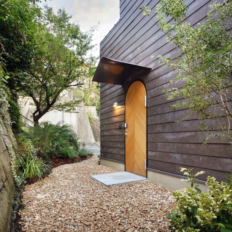 Exterior view of a modern home with dark siding, light wood doors, and clean landscaping.