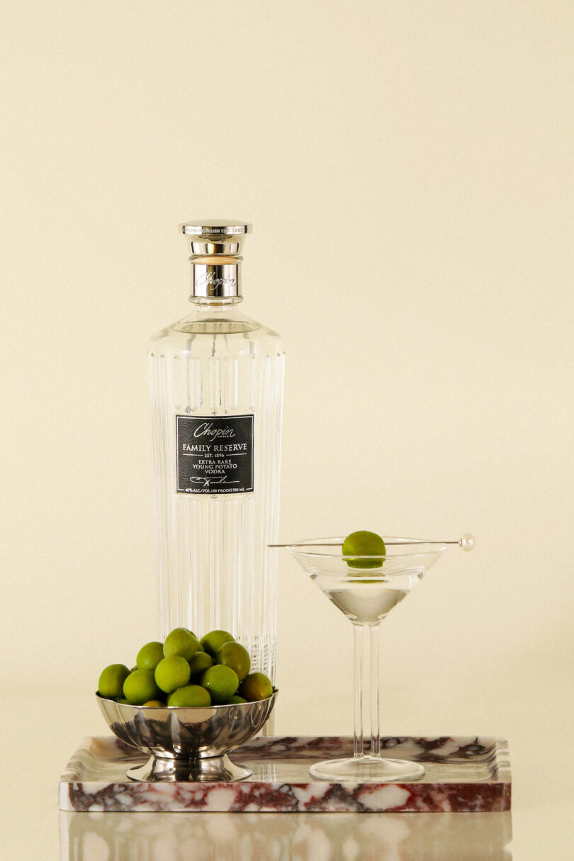 A vodka bottle and a martini glass arranged with a silver bowl of olives on a white marble slab.