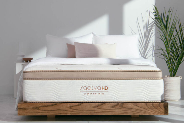 A luxury mattress on a wooden platform bed in a bright, modern bedroom with plants and natural lighting.