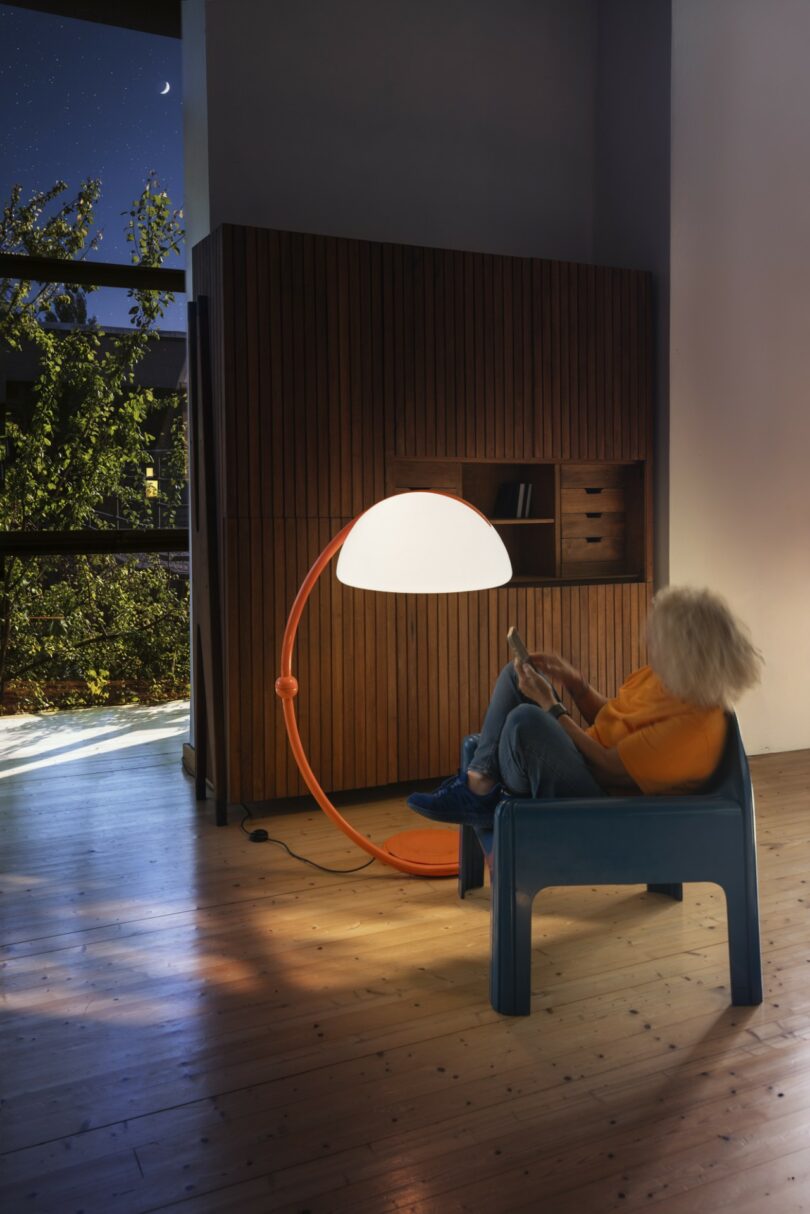 A person reading a book in a blue chair illuminated by an orange Serpente floor lamp in a cozy interior.