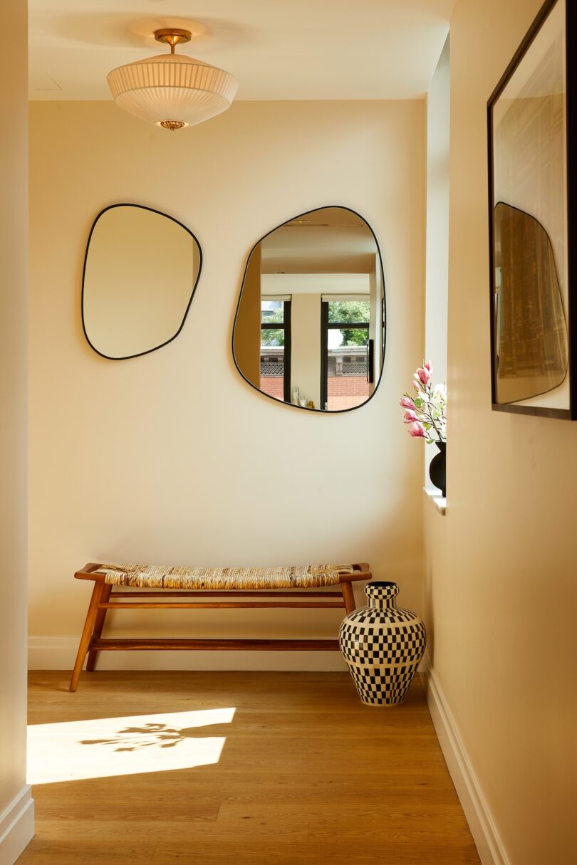 A bright hallway featuring two asymmetrical mirrors, a woven bench, and light wood floors.