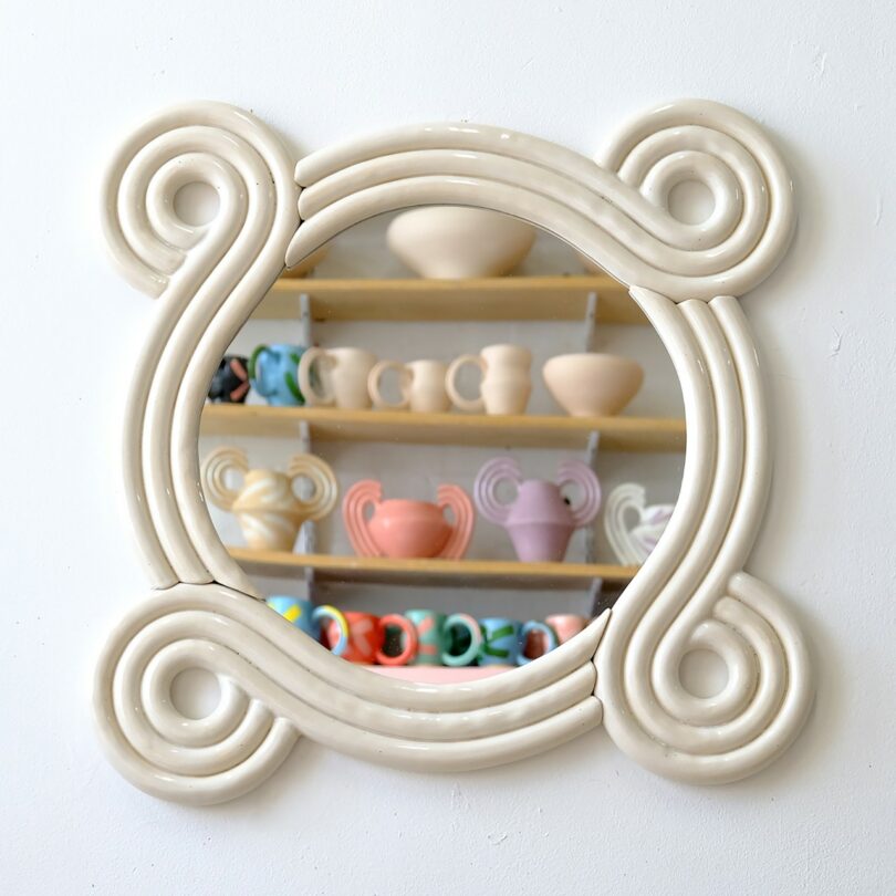 A round white wavy mirror reflecting shelves of colorful ceramic cups and vases in a studio.