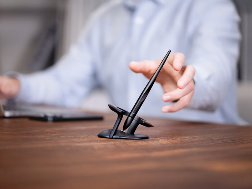 A person's hand reaching towards a black Novium Hoverpen balanced in its sculptural stand on a wooden desk.