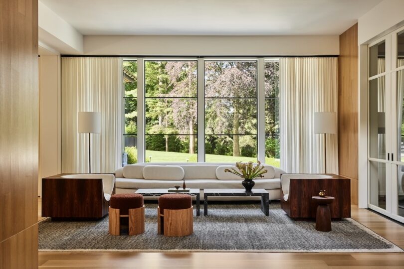 Modern living room with floor-to-ceiling windows overlooking lush greenery, featuring neutral-toned furniture.